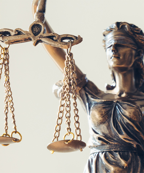 The image features a close-up view of a statue of Lady Justice, focusing on her raised hand holding the scales of justice. The statue is crafted in bronze with intricate detailing, highlighting the symbolic scales that are evenly balanced.